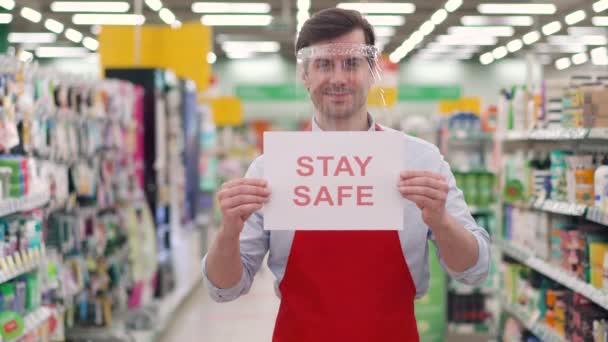 Щасливий роздрібний робітник у червоному фартусі з пластиковим щитом на обличчі стоїть у супермаркеті з паперовою маркою "Залишайся в безпеці". Роботодавець, який працює в магазині під час епідемії covid-19, концепт пандемії коронавірусу — стокове відео