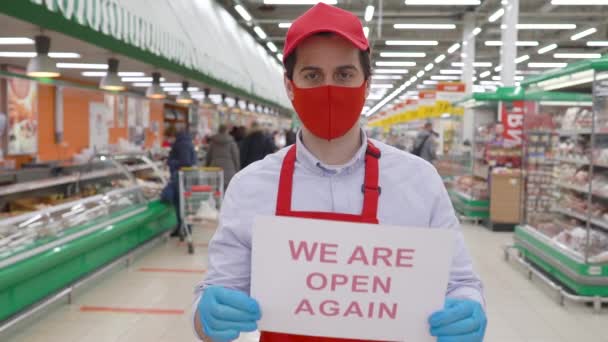 Vendedor hombre en máscara roja, guantes y uniforme de pie en el supermercado sosteniendo un papel etiquetado estamos abiertos de nuevo. Empleado trabajando en tienda de comestibles durante epidemia covid-19, concepto pandémico coronavirus — Vídeo de stock
