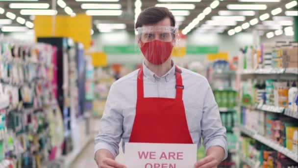 Vedoucí prodeje v červené masce a plastový štít stojí v supermarketu s papírem s nápisem Jsme opět otevřeni. Zaměstnanec pracující v obchodě během epidemie covid-19, koronavirus pandemické koncepce — Stock video