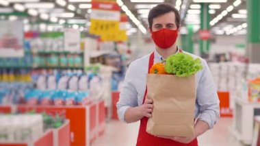 Yüz maskeli kurye ve kırmızı üniformalı süpermarkette elinde sebzelerle duruyor. Salgın covid-19, koronavirüs salgını, çevrimiçi gıda alışverişi sırasında markette çalışan kurye.