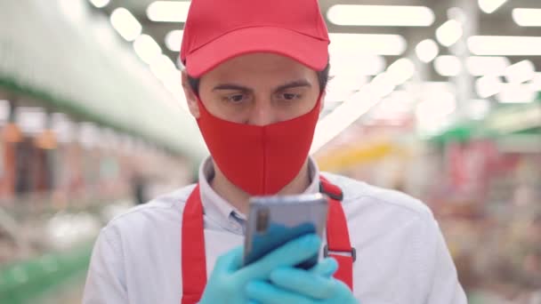 Trabalhador de varejo masculino em máscara vermelha e luvas em pé no supermercado e usando smartphone, tomando ordens de comércio eletrônico on-line. Vendas em mercearia durante epidemia de covid-19, pandemia de coronavírus — Vídeo de Stock