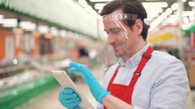 Plastik bir yüz kalkanındaki erkek perakende işçisi süpermarkette dijital tablet kullanarak çevrimiçi ecommerce siparişleri alıyor. Salgın covid-19, koronavirüs salgını sırasında markette çalışan satışlar.