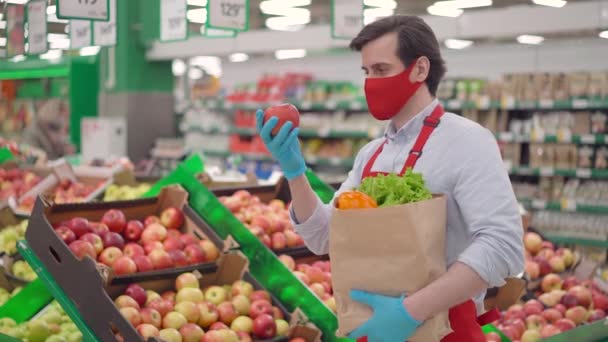 Repartidor masculino con máscara facial roja de pie en la tienda de comestibles seleccionando manzanas frescas. Mensajero trabajando en el servicio de entrega de supermercados durante epidemia covid-19, pandemia de coronavirus, compras de alimentos en línea — Vídeo de stock