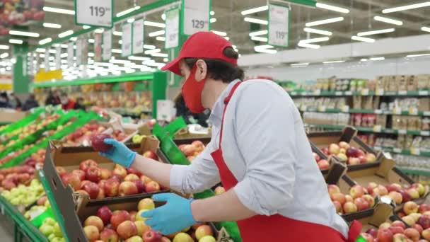 Repartidor masculino con máscara facial roja de pie en la tienda de comestibles seleccionando manzanas frescas. Mensajero trabajando en el servicio de entrega de supermercados durante epidemia covid-19, pandemia de coronavirus, compras de alimentos en línea — Vídeo de stock
