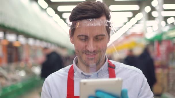 Férfi eladó műanyag arc pajzs áll a szupermarketben digitális tabletta, figyelembe online e-kereskedelmi megrendelések. Eladó férfi dolgozik élelmiszerbolt idején járvány covid-19, coronavirus pandemic — Stock videók