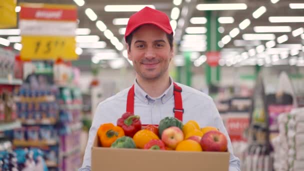 Feliz jovem entregador ou mensageiro vestindo uniforme vermelho segurando caixa de papelão com legumes frescos em pé na loja do supermercado. Rápido expresso e seguro entrega de alimentos compras on-line, conceito de comércio eletrônico — Vídeo de Stock