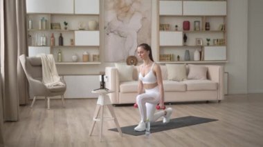 Sportif genç bir kadın dambıllarla online egzersiz yapıyor. Aktif bir kız akıllı telefonuyla spor antrenmanından zevk alıyor yoga minderinde gerinerek video izliyor. Spor ve eğlence konsepti.