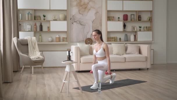 Sportieve jonge vrouw doet online training met halters. Actief meisje genieten van sport fitness training op smartphone thuis, stretching op yoga mat kijken video. Sport en recreatie. — Stockvideo
