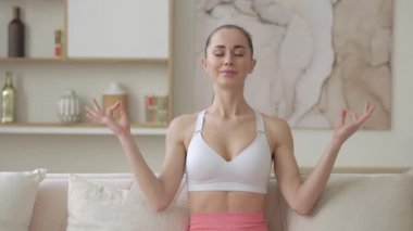 Düşünceli, genç, melez bir kadın mudra jesti yapıyor. Evde rahat bir kanepede lotus pozisyonunda oturuyor. Huzurlu bir milenyum kızı derin meditasyon yapıyor, yoga egzersizleri yapıyor.