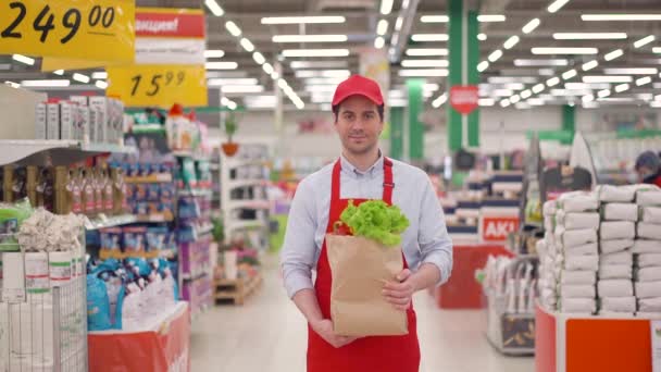 Hombre joven vendedor caucásico guapo en uniforme rojo sosteniendo paquete de papel artesanal con verduras frescas de pie en el supermercado. Compras en línea rápidas y seguras de entrega de alimentos, concepto de comercio electrónico — Vídeos de Stock
