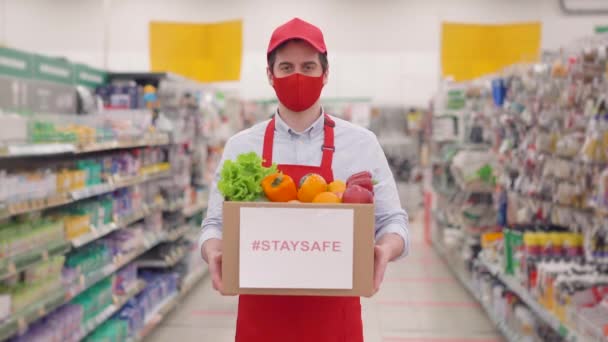 Mężczyzna kurier sprzedawca człowiek w czerwonym mundurze i maski do twarzy trzymając pudełko kartonowe ze świeżych warzyw stojących w supermarkecie. Szybka ekspresowa bezpieczna dostawa podczas pandemii 19, koncepcja zakupów online — Wideo stockowe