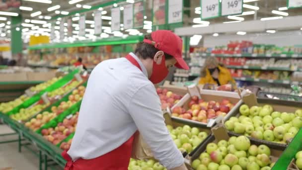 Vendedor de mascarilla roja de pie en la tienda de comestibles seleccionando manzanas frescas. Mensajero trabajando en el servicio de entrega de supermercados durante epidemia covid-19, pandemia de coronavirus, compras de alimentos en línea — Vídeo de stock