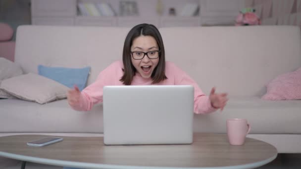 Excited asian girl winner looks at laptop computer, screaming yes celebrates mobile online bid game win victory success siting on sofa at home. Mixed race lady using app celebrating success concept — Stock Video