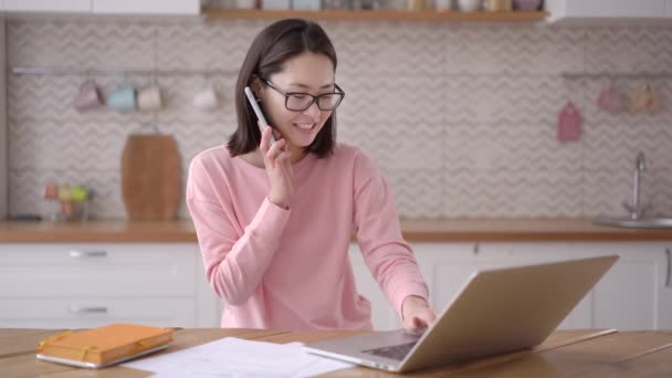 Sonriente joven mixta mujer de negocios asiática hablando por teléfono usando el ordenador portátil sentarse en el escritorio de la oficina en casa, feliz cliente femenino hacer una llamada móvil confirmando el sitio web en línea compras entrega de pedidos concepto — Vídeos de Stock