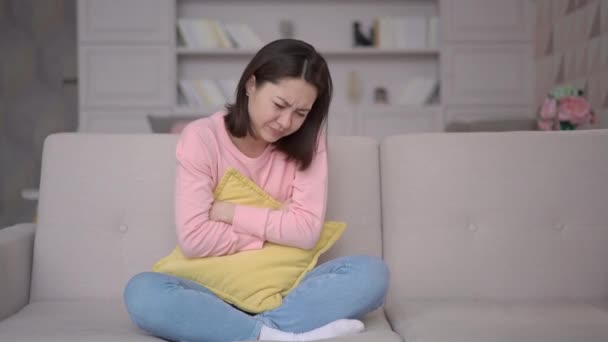 Stressed multi ethnic woman sitting on sofa, hugging pillow embracing belly, suffering from stomachache. Asian girl having menstrual painful feelings, gastritis, diarrhea and painful periods concept — Stock Video