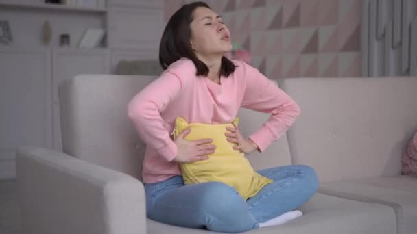 Stresowana mieszana kobieta rasy siedząca na kanapie, przytulająca poduszkę i obejmująca brzuch, cierpiąca na ból brzucha. Azjatka o miesiączki bolesne uczucia, zapalenie błony śluzowej żołądka, biegunka i bolesne pojęcia okresów — Wideo stockowe