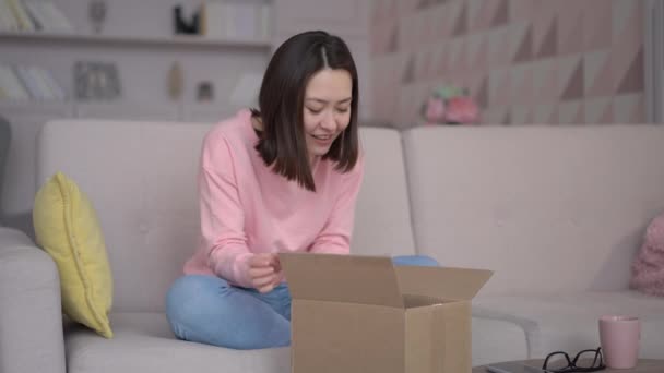 Ung multietnisk kvinne som åpner pakkeboks hjemme. En begeistret asiatisk shopper som åpner motekjøp på sofaen. Forbrukervennlig metode for utpakking av postforsendelse – stockvideo
