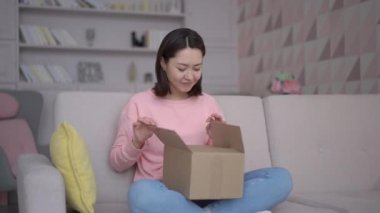 Young asian female customer opening cardboard box at home. Excited multi ethnic girl shopper unboxing fashion purchase sitting on sofa. Satisfied consumer unpacking postal shipping delivery parcel
