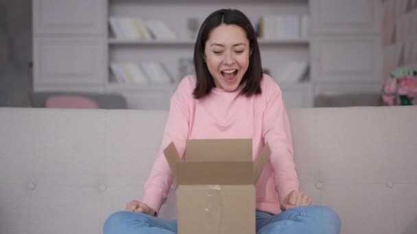 Счастливая миллениалка сидит дома на диване, распаковывает картонную коробку с интернет-заказом, совершает покупки онлайн, улыбается юной клиентке, распаковывает почтовый ящик с концепцией службы доставки товаров — стоковое видео