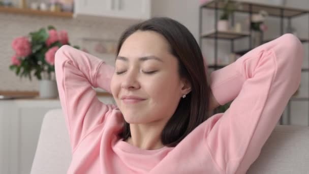 Smiling multi ethnic asian woman resting on sofa holding hands behind head eyes closed take deep breath of fresh air dream lounge on couch at home feel no stress concept enjoy comfort welfare at home — Stock Video