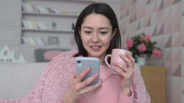 Wieloetniczna młoda azjatycka dama korzystająca ze smartfona surfująca po mediach społecznościowych, sprawdzająca wiadomości, grająca w gry mobilne lub wysyłająca SMS-y na sofie. Millennial kobieta spędza czas w domu z gadżetem — Wideo stockowe