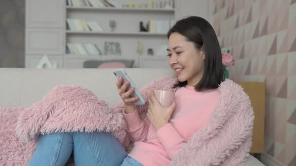 Die multiethnische junge Asiatin surft mit ihrem Smartphone in den sozialen Medien, checkt Nachrichten, spielt Handyspiele oder schreibt auf dem Sofa sitzend Nachrichten. Millennial-Frauen verbringen Zeit zu Hause mit Gadget — Stockvideo