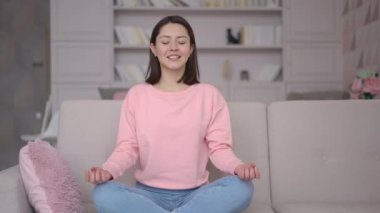 Karışık ırktan Asyalı bir kadın oturma odasındaki rahat koltukta bağdaş kurarak oturur. Gözler kapalı, iç ahenk ve dengeyi hisseder. Huzurlu zihin ve zen için yoga nefesini geliştirir.