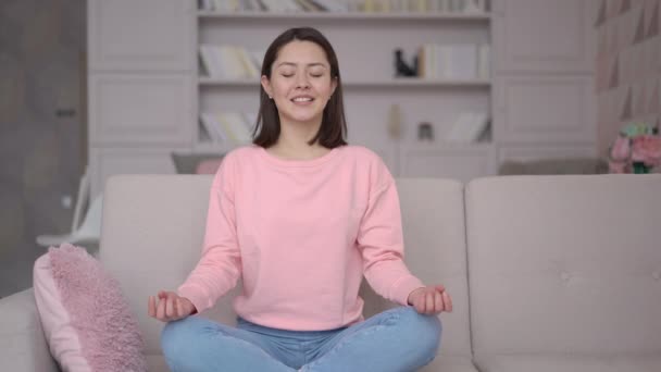 Blandad ras asiatisk kvinna sitta över ben på bekväm soffa i vardagsrummet slutna ögon praxis känns inre harmoni och balans, förbättra självmedveten, utövar yoga andning för fridfullt sinne och zen — Stockvideo