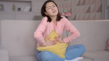 Stresli çok ırklı kadın koltukta oturuyor, yastığa sarılıyor karnına sarılıyor, karın ağrısı çekiyor. Adet sancısı çeken Asyalı bir kız, mide iltihabı, ishal ve acı dolu dönem konsepti.