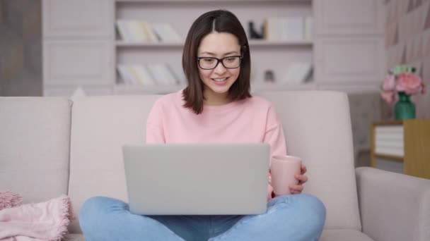 Mujer asiática sentada en un sofá usando una computadora portátil mirando el mensaje de escritura de pantalla, señora multiétnica charlando en la computadora navegando por Internet, estudiando redes sociales, trabajando en línea en casa — Vídeos de Stock