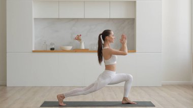 Beyaz spor giyimli, pantalon ve bluz giyen, oturma odasında spor minderleri, sağlık hizmetleri ve ev konseptinde zihinsel terapi üzerine yoga egzersizleri yapan çekici, etnik kökenli genç bir kadın.