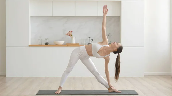 Beyaz spor giyimli, pantalon ve bluz giyen, oturma odasında spor minderleri, sağlık hizmetleri ve ev konseptinde zihinsel terapi üzerine yoga egzersizleri yapan çekici, etnik kökenli genç bir kadın.