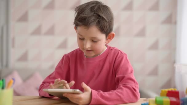 Забавный школьник с планшетным компьютером, сидит дома один, позитивный ребенок смотрит на экран, играет в видеоигры, смотрит мультики, просматривает приложения, веселится с устройством — стоковое видео