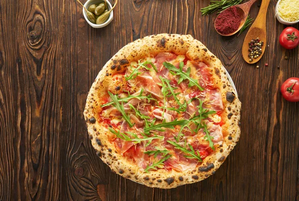 Oryginalna włoska neapolitańska pizza z szynką parmeńską i świeżą rukolą na drewnianym tle ze składnikami. Fast food menu koncepcja jedzenia, widok z góry — Zdjęcie stockowe