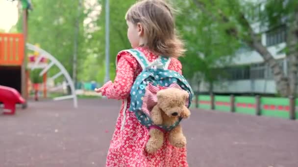 Мила дівчинка-малятко, що йде по дорозі на міський майданчик з іграшковим ведмедем у рюкзаку на спині. Маленька дитина розважається на відкритому повітрі, догляд за дітьми та активна концепція дошкільника — стокове відео