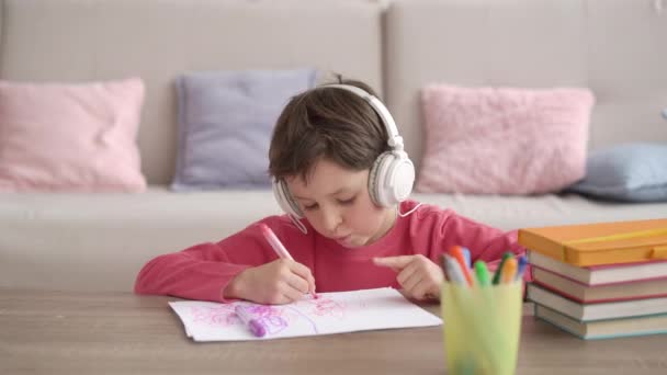 Cute little schoolboy dziecko w słuchawkach słuchać muzyki, rysunek obraz z kolorowych ołówków w papierowym albumie sam w domu. Kaukaski mały dzieciak kolorowanki malarstwo, ciesząc się czas hobby w salonie. — Wideo stockowe
