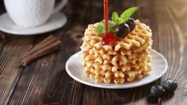 Böğürtlenli ve beyaz tabakta naneli waffle ile süslenmiş taze pişmiş Belçika wafflelarının üzerine çilek şurubu dökülüyor. Tahta masada ya da kırsal arka planda kahvaltı için nefis ev yapımı hamur işi.