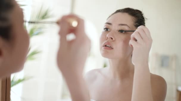 Ελκυστική νεαρή πολυεθνική γυναίκα που κοιτάζει στον καθρέφτη στο μπάνιο του σπιτιού βάφει τις βλεφαρίδες με μαύρη πυκνότερη μάσκαρα όγκου, αρκετά χιλιετή μικτή φυλή ασιατικό θηλυκό κάνει πρωινές διαδικασίες μακιγιάζ. — Αρχείο Βίντεο