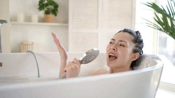 Vzrušená mladá asijská žena zpívající ve vaně pomocí sprchové hlavice. Šťastné tisícileté multietnické ženy baví těšit každodenní péči v koupelně, kosmetické terapie a zdravý životní styl koncept — Stock fotografie