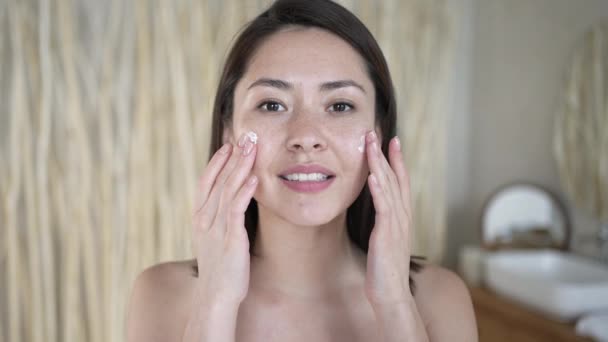 Aantrekkelijke Aziatische vrouw toe te passen huidverzorgingscrème op gezicht look in badkamer spiegel thuis. Gelukkig jong gemengd ras dame zet hydraterende liftende gezichtscrème, gezonde huidverzorging en lichaamsverzorging concept. — Stockvideo