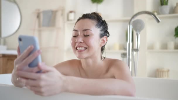 Atractiva joven mujer asiática de raza mixta haciendo videollamada usando un teléfono inteligente tumbado en la bañera. Millennial chica en baño con teléfono móvil que tiene webcam video conversación disfrutar del cuidado del cuerpo diario en el baño — Vídeos de Stock