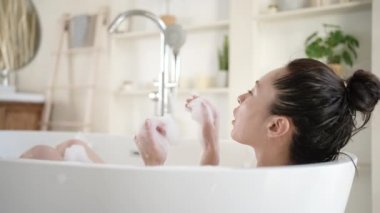 Çekici genç Asyalı kadın küvette köpükle oynuyor. Mutlu milenyum çok ırklı kadınlar banyoda günlük vücut bakımının, güzellik tedavisinin ve sağlıklı yaşam tarzı konseptinin tadını çıkarıyor.