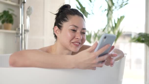 Attractive multi ethnic asian female making video call using smartphone lying in bathtub. Millennial asian girl with mobile phone having webcam video conversation enjoy daily bodycare in bathroom — Stock Video