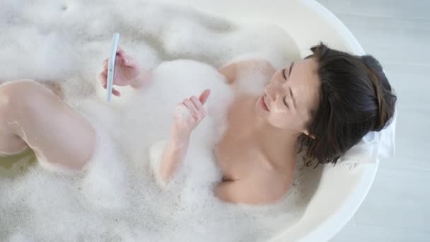 Attractive young mixed race asian woman making video call using smartphone lying in bathtub. Millennial girl in bath with mobile phone having webcam video conversation enjoy daily bodycare in bathroom — Stock Video