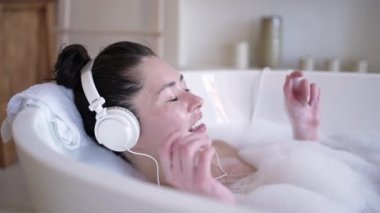 Küvette gözleri kapalı şarkı söyleyen kulaklıklı çekici Asyalı bir kadın. Çok ırklı milenyum kızı rahat müzik dinliyor banyoda dinleniyor, tedavi görüyor, sağlıklı yaşam tarzı kavramları var.