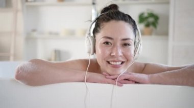 Küvette şarkı söyleyen kulaklıklı çekici, gülümseyen, melez Asyalı bir kadın. Çok ırklı milenyum kızı banyoda rahat müzik dinliyor, tedavi görüyor sağlıklı yaşam tarzı konsepti.