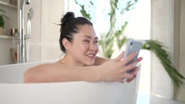 Çekici, çok kültürlü Asyalı kadın küvette uzanan akıllı telefonla video görüşmesi yapıyor. Cep telefonlu genç bir kadın web kamerası görüntülü konuşma yaparak banyoda günlük vücut bakımının keyfini çıkarıyor.