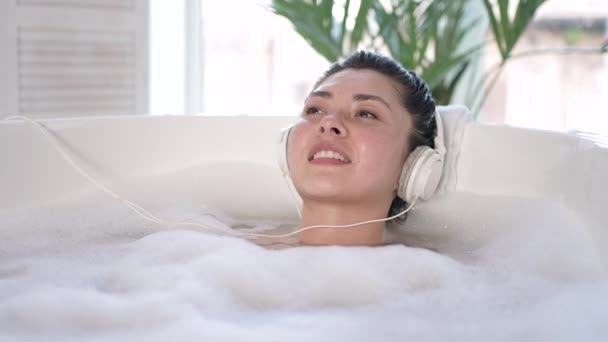 Attraktiv ung sensuell asiatisk brunett kvinna med hörlurar njuter av ett varmt bad. Multi etnisk tusenårig flicka lyssnar chill musik avkopplande i badrummet, behandling terapi, hälsosam livsstil koncept — Stockvideo