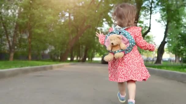 Bayi perempuan kecil yang lucu berjalan di jalan di taman bermain kota dengan mainan beruang di ransel di punggungnya. Anak kecil bersenang-senang di luar ruangan, tempat penitipan anak dan konsep aktif pra-SD — Stok Video