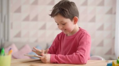 Komik okul çocuğu tablet bilgisayar kullanıyor, evde tek başına oturuyor, pozitif çocuk Gadjet ekranına bakıyor, video oyunu oynuyor, çizgi film izliyor, uygulamaları tarıyor, cihazla eğleniyor.